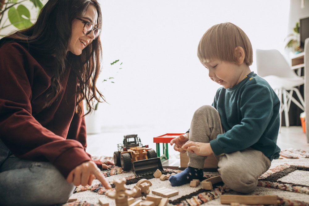 Hlídání dětí jako jedna z možností podnikání - obrázek zobrazující ženu, která si hraje s dítětem, blonďatým chlapcem, s dřevěnými hračkami.